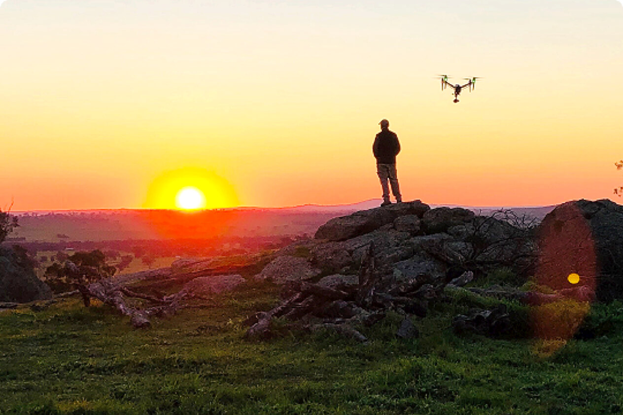 Man standing on rock watching sunset over grassy landscape, drone in air behind him